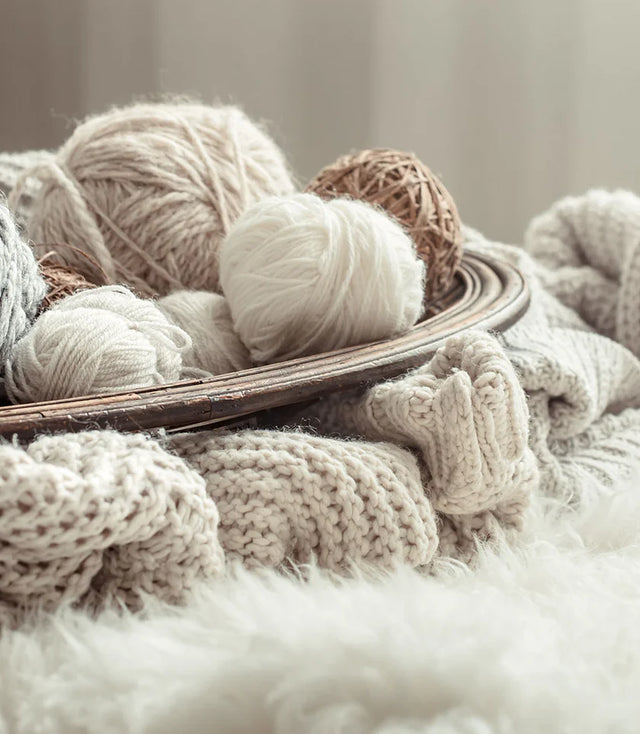 Gomitoli di lana naturale in diverse tonalità di bianco, beige e crema, disposti su un vassoio con morbide coperte lavorate a maglia.