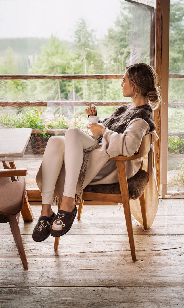 Donna seduta su una sedia di legno in una baita rilassato mentre prende il caffè con indosso le pantofole della linea flair di haflinger coffee time marroni