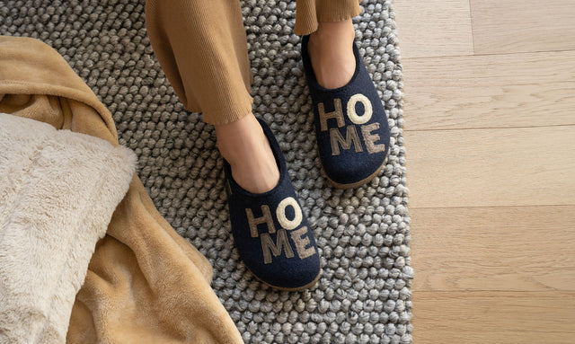 primo piano di piedi donna poggiati per terra su un tappeto. Indossa delle ciabatte Haflinger di colore blu con scritto sopra "Home". il modello si chiama Home ed è unisex.