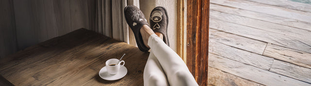 Primo piano di pantofole haflinger in lana con ricamo coffee time indossate da donna con le gambe appoggiate al tavolo