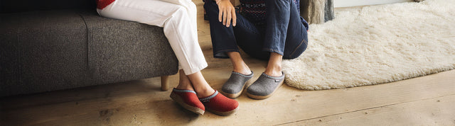 Primo piano di due paia di pantofole sabot haflinger franzl, un paio rosso indossato da donna seduta su una poltrona e un paio grigio indossato da uomo seduto su un tappeto vicino alla donna