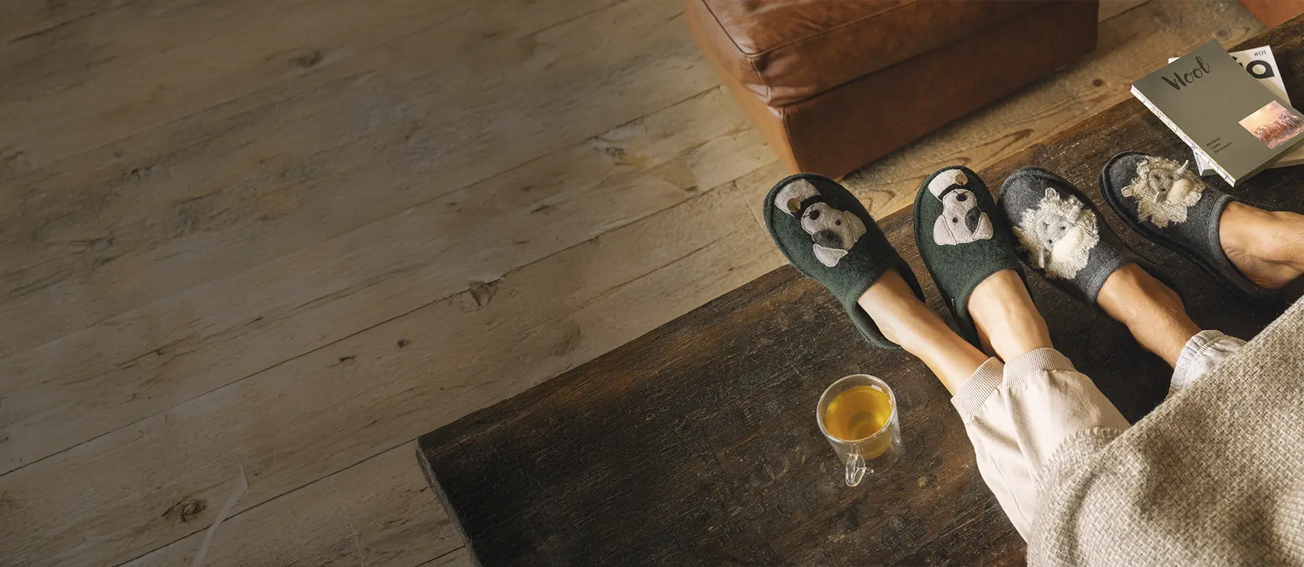 Primo piano di pantofole haflinger in lana: un paio indossato da uomo con decorazione pecora del modello lamby e un paio con decorazione cane theo indossato da donna. Piedi appoggiati a un table coffee